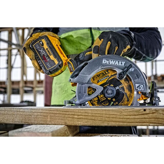 En person håller i en DeWalt cirkelsåg med ett 18V batteri, placerad på en träbänk. Bakom syns byggnadsställningar.