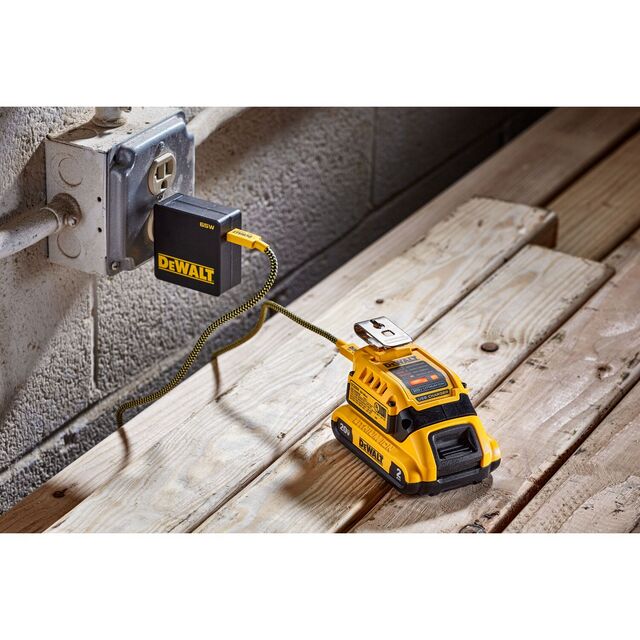 Gult batteri från DeWalt laddas i ett vägguttag, placerat på träplankor mot en betongvägg.
