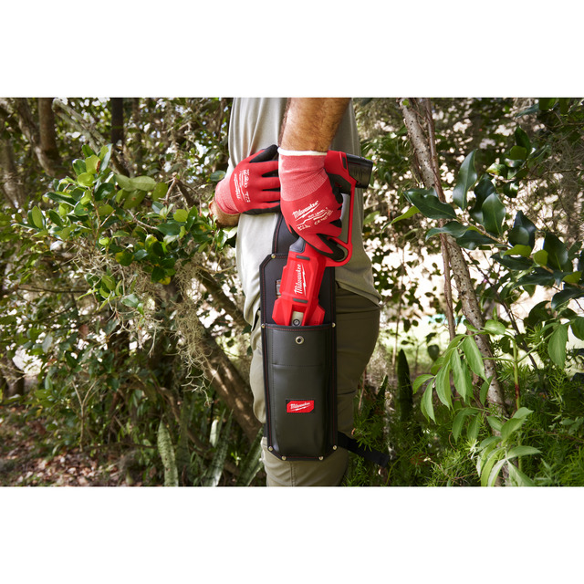 Person wearing red gloves holding a tool in a black holster in a garden setting.