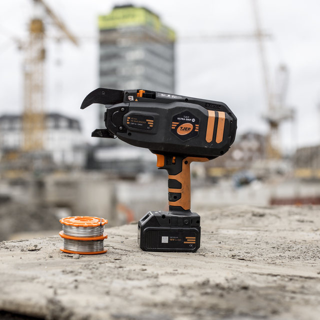 Verktyg för byggarbetsplats, orange-svart, står på betongyta bredvid rulle med ståltråd. Bakgrund med kranar och byggnader.