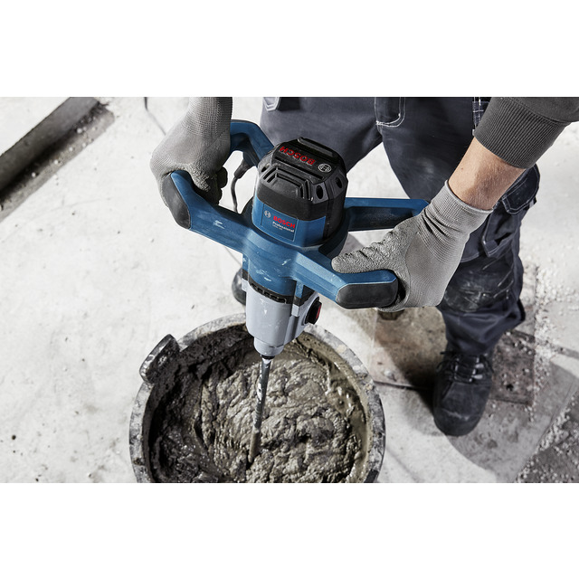 En person med handskar håller en elektrisk blandare som rör om cement i en hink.