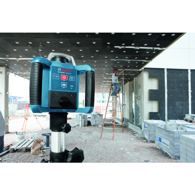 Automatisk rotationslaser på stativ, byggnadsarbetare på stege, oavslutat innertak och byggmaterial i bakgrunden.