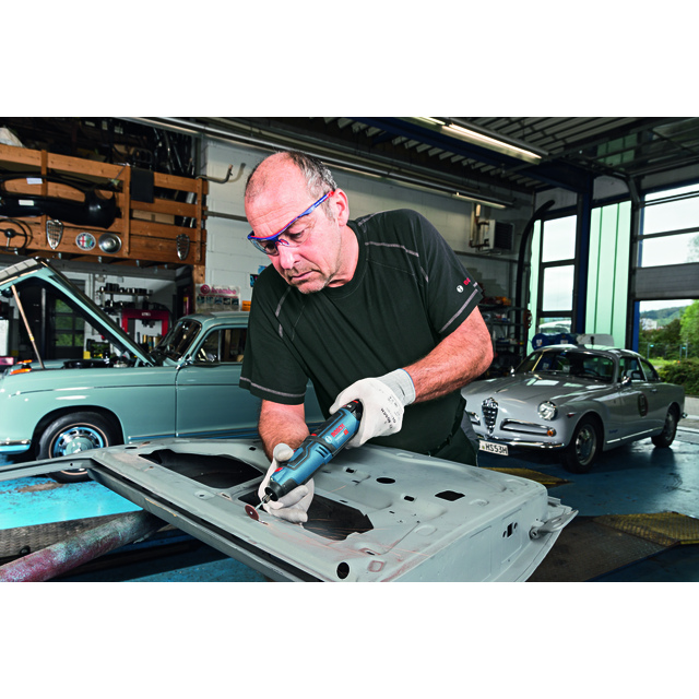 Man in protective glasses working on metal sheet in garage with classic cars in background.