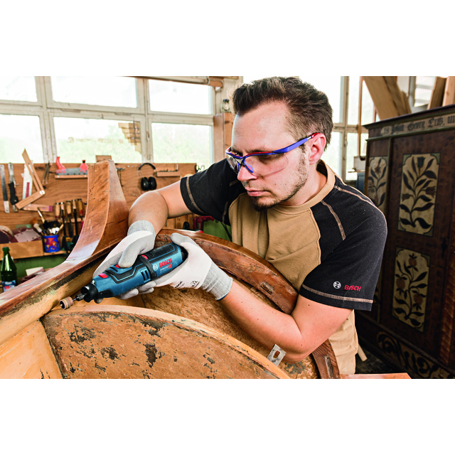 Man in workshop using a Bosch rotary tool on wooden furniture, wearing gloves and safety glasses.