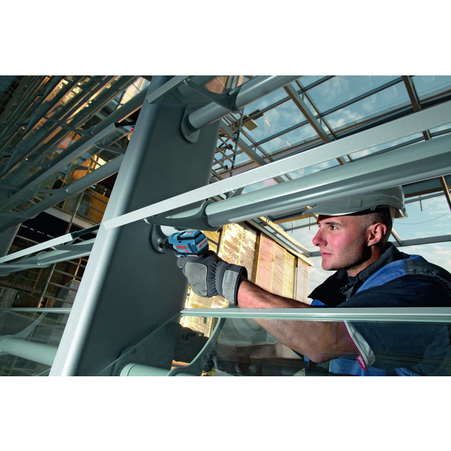 Person med hjälm och arbetshandskar använder en elektrisk skruvdragare på en metallstruktur inomhus.