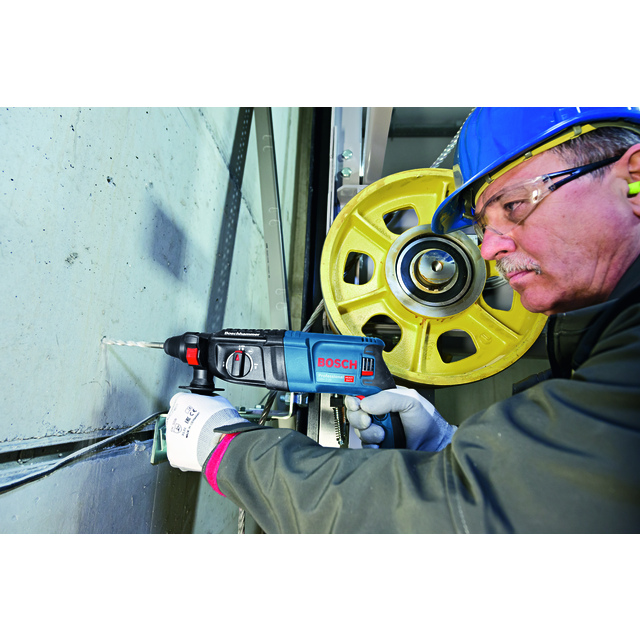 Man in safety gear using a Bosch drill on a concrete wall, with a large yellow pulley wheel in the background.