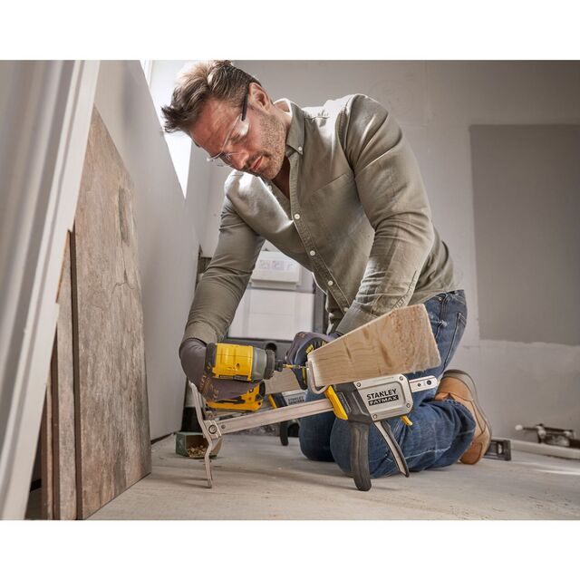 Man using a Stanley clamp and electric drill on a wooden piece, wearing safety glasses and gloves in an indoor setting.
