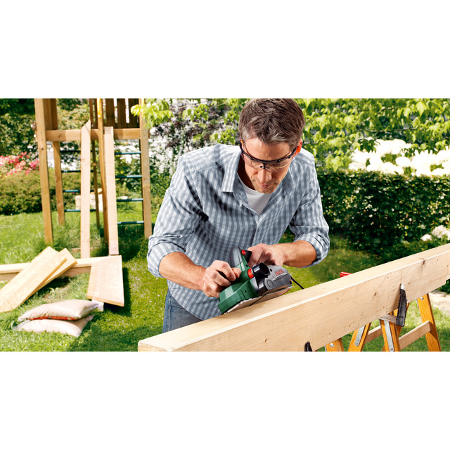 Man wearing safety glasses using an electric planer on a wooden beam in a garden setting.