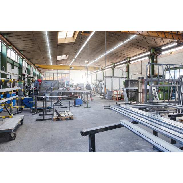 Verkstad med metallstänger, verktyg och industriutrustning. Takbelysning och stora fönster syns i bakgrunden.