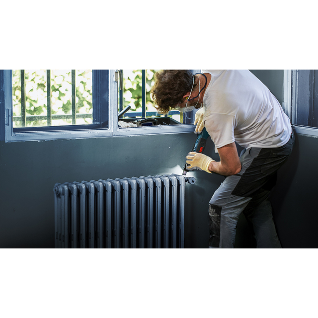 Person i arbetshandskar och skyddsöronsnäckor arbetar med verktyg vid en radiator nära ett fönster.