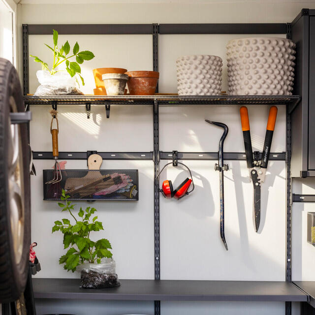Hylla i garage med växter, krukor, trädgårdsverktyg, hörselskydd och upphängd borste.