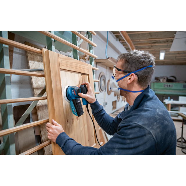Person slipar trädörr med elektrisk slipmaskin inomhus. Bär skyddsglasögon och ansiktsmask.